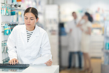 Positive young female pharmacist standing at the desk using keyboard in chemist's shop