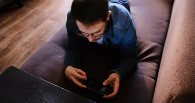 Immerse into gaming with a close-up of a man in a white tee and jeans playing a handheld console lying on a brown couch. Ideal for lifestyle gaming-themed videos. selective focus cat walking in shoot
