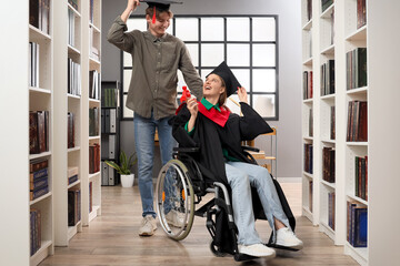 Female graduate in wheelchair with diploma and her classmate at library