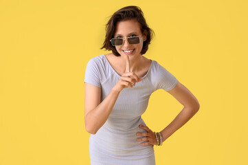 Pretty young woman with sunglasses showing silence gesture on yellow background