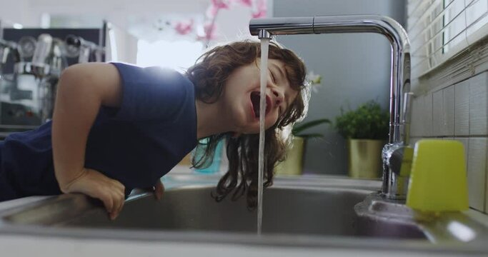 Cute boy with curly hair leans over a kitchen sink to drink water straight from the tap, with daylight illuminating the scene.