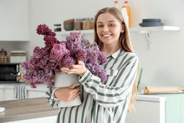 Young woman holding vase of lilacs near counter in kitchen