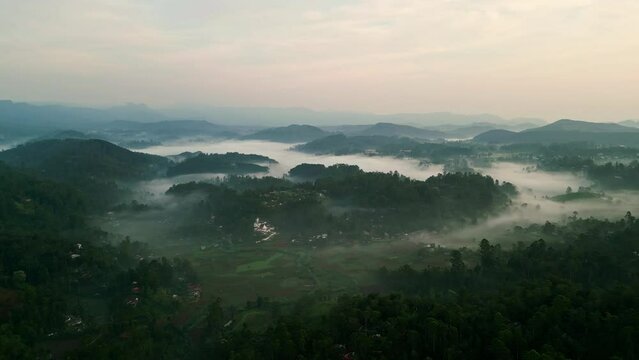 Aerial drone sweeps over misty Haputale mountains in Sri Lanka at dawn. Clouds envelop green highlands, terraced tea plantations. Sunrise illuminates serene landscape, inviting exploration, adventure.