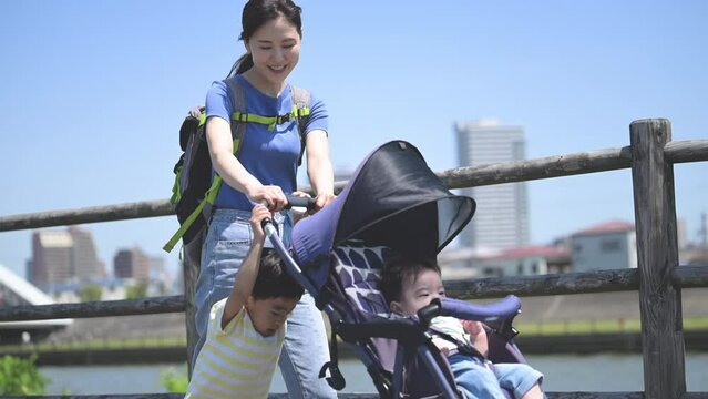 晴天の川沿いの公園でベビーカーで散歩するママと兄弟の親子(3人家族)の上半身　春夏のお出かけやママの通勤などのイメージのスロー動画