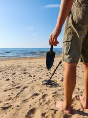 A man is standing on the beach with a metal detector in his hand, leisurely searching for metal...