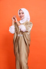 Young asian Muslim woman in hijab isolated orange background. standing at an angle to the left, cheerful, hands together to the side, wearing an Islamic caftan. Half body portrait for fashion