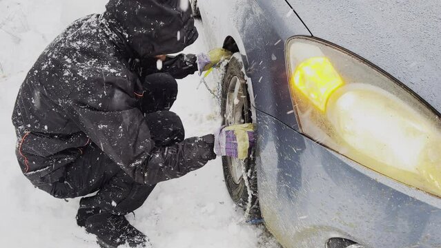 A man crouches beside a car, braving a snowstorm to fit snow chains onto the tire for safer travel in winter conditions.