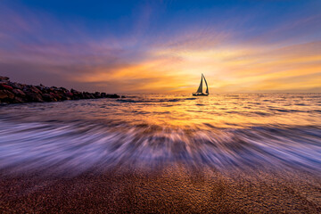 Vibrant sunset over the ocean with a sailboat on the horizon and gentle waves in the foreground