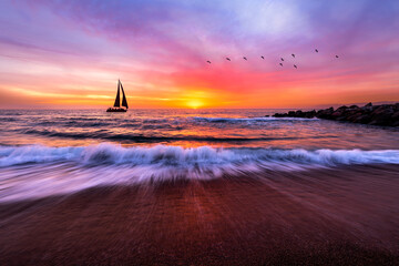 Sailboat drifts by under a vibrant sunset sky, as birds fly and waves lap the shore