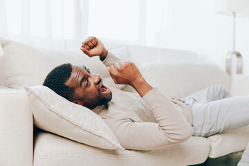 Stressed African American man with a headache sitting alone on a couch at home He looks tired, sick, and frustrated, showing signs of mental and physical pain The background is a living room, where he