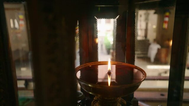 Oil scent lamp burns in Buddhist temple, flame flickers with serene ambiance in spiritual setting, Kandy, Sri Lanka. Devotees walk, worship in blurred background peaceful meditation atmosphere.