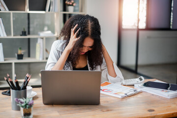 Businesswoman feeling stressed while working on her laptop, surrounded by documents and a calculator, indicating a challenging work situation.
