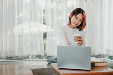 Casual young woman taking a break with a coffee cup, seated comfortably at her laptop in a well-lit cozy room, embodying a relaxed work-from-home atmosphere.