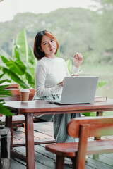 Young woman sits thoughtfully at a wooden table with her laptop, surrounded by the lush greenery of an outdoor study area.