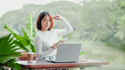 Contemplative young woman studies outdoors with a laptop and notebook, surrounded by lush greenery and the tranquility of nature.