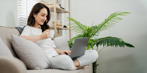 Comfortable young woman enjoying a hot drink while working on her laptop in a cozy home environment.