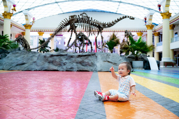 Asian little child looks at a replica skeleton of an ancient dinosaur in an paleontology museum....