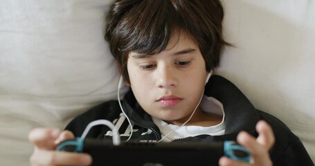 Cute young boy wearing headphones playing a game on a handheld gaming console, comfortably reclining on a couch indoors, daylight.