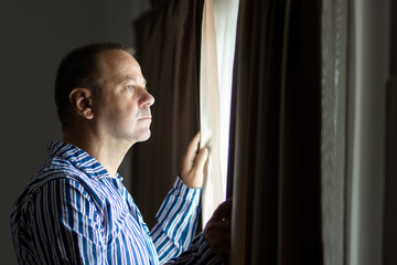 A man in striped pajamas looks out of a curtained window