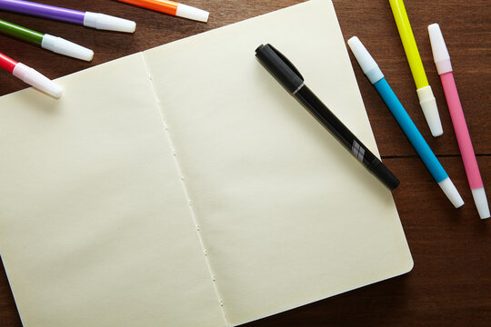 an empty sketchbook opened with a black marker and a number of coloured markers on the wooden table