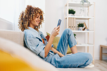 Relaxed Woman Sitting on Sofa, Holding Phone and Smiling, Enjoying Online Games and Chatting in...