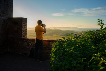 A photographer in a yellow shirt captures the sunset from an ancient wall, with a picturesque...
