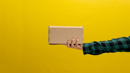 A hand holding a cardboard box against a yellow background, illustrating the concept of shipping,...