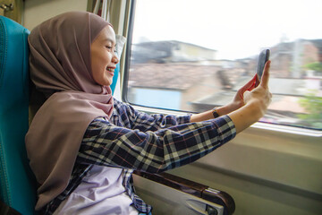 happy asian muslim woman holding her mobile phone while sitting beside big window train wagon. traveling concept