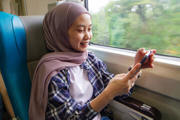 happy asian muslim woman holding her mobile phone while sitting beside big window train wagon. traveling concept