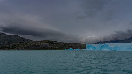 A wall of blue ice stretches over a turquoise glacial lake. Coastal mountains against the sky and clouds. Canoes with tourists sail to the glacier. Perito Moreno. El Calafate. Argentina.Lago Argentino