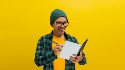 A diligent young Asian student, wearing glasses, a beanie hat, and casual clothes, is captured taking notes while holding a notebook and pen. Studying, learning languages, and preparing for exams.