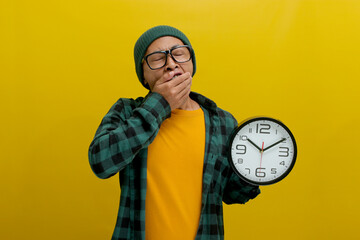 A sleepy young Asian man, dressed in a beanie hat and casual shirt, yawns while holding a clock,...