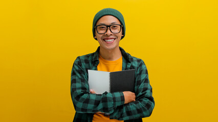 A young Asian student, wearing eyeglasses, a beanie hat, and casual clothes, stands against a yellow background, holding books against his chest.