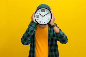 Asian man, dressed in beanie hat and casual shirt, creatively uses a clock to cover his head or...