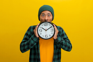 An Asian man, sporting a beanie hat and casual shirt, holds a clock in front of his face, looking...