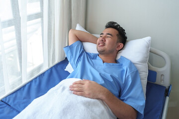 A smiling Asian man lies in a hospital bed, relaxed, showing a fighting pose after receiving...