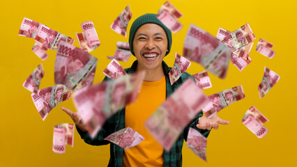 Cheerful Asian man in a beanie and casual clothes celebrates success with money banknotes flying in...