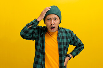 Shocked and frightened young Asian man, dressed in a beanie hat and casual shirt, reacts to something alarming while standing against a yellow background