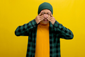 Young Asian man in a beanie hat and casual shirt is rubbing his swollen eyes, possibly indicating...