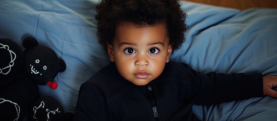 An adorable African American baby boy dressed in black is seen top view as he plays with toys and relaxes on a bed at home in a bedroom He can be seen holding a rattle and looking directly at the cam - Powered by Adobe