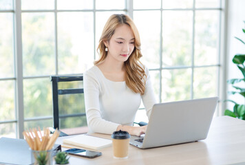 Asian pretty young happy professional businesswoman employee sitting smiling professional at desk working on laptop in office. Attractive female thinking worker using computer at workplace.