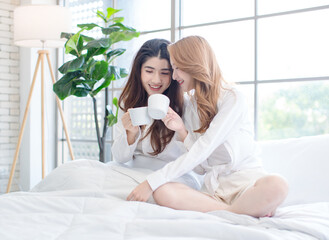 Two beautiful Asian female girls drinking coffee in a bedroom, Cheerful young woman smiling, happy sitting on the bed, having a conversation, drinking tea, weekend breakfast lifestyle at home