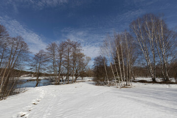 Tranquil winter landscape with snow-covered ground, bare trees, and a partially frozen body of water reflecting the blue sky with scattered clouds. Footprints lead through the snow