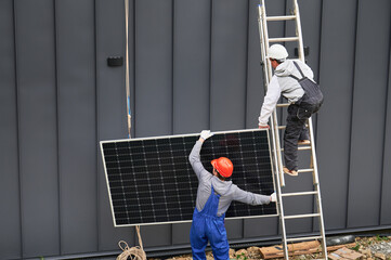 Men builders installing solar panel system on roof of house. Electricians in helmets lifting up...
