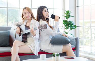 Two asian couple beautiful woman playing acoustic guitar, sing folk song together sitting on sofa...