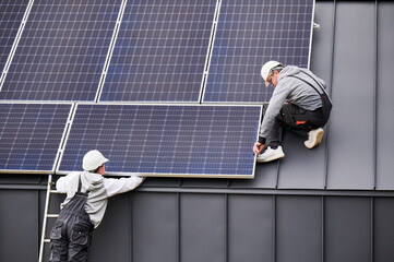 Technicians connecting cables while installing photovoltaic solar panels on roof of house. Workers...
