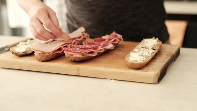 Caucasian male placing sliced turkey ham on a halved brown baguette in the kitchen on the countertop.