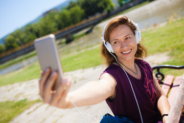 A cheerful young woman listens to music with white headphones and takes a selfie on a sunny day in...