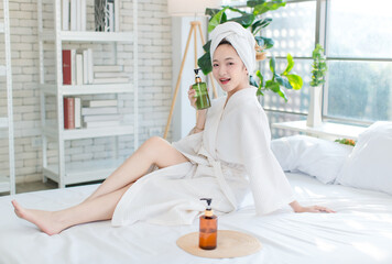 Asian happy beautiful young woman smiling wearing a towel and a white bathrobe sitting relaxed enjoy having fun, on the bed in bedroom spending time together at home, spa concept.