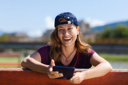A joyful young woman wearing a baseball cap sitting outdoors, laughing while looking at her smartphone on a sunny day near a river.